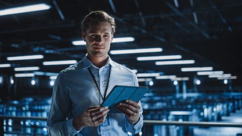 Ein junger Mann steht mit einem Tablet vor einer technischen Kulisse. Er hat kurze, braun-blonde Haare und trägt ein hellblaues Hemd. Um den Hals trägt er einen Bediensteten-Ausweis.